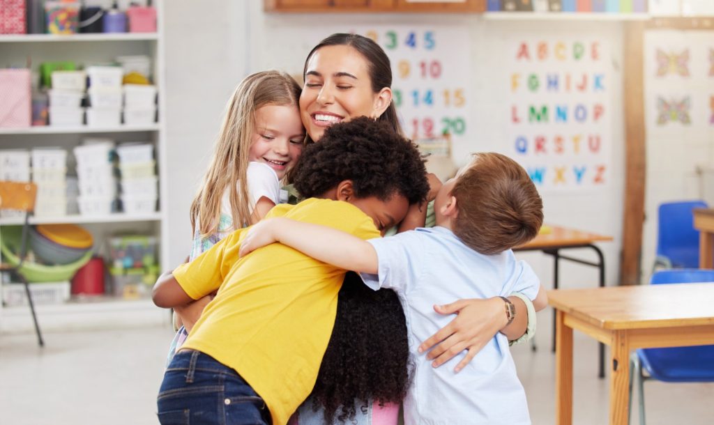 Care, teacher with school children hug and in a classroom with a lens flare. Support or love, happiness or teaching and happy or cheerful woman with kindergarten kids hugging together in a class