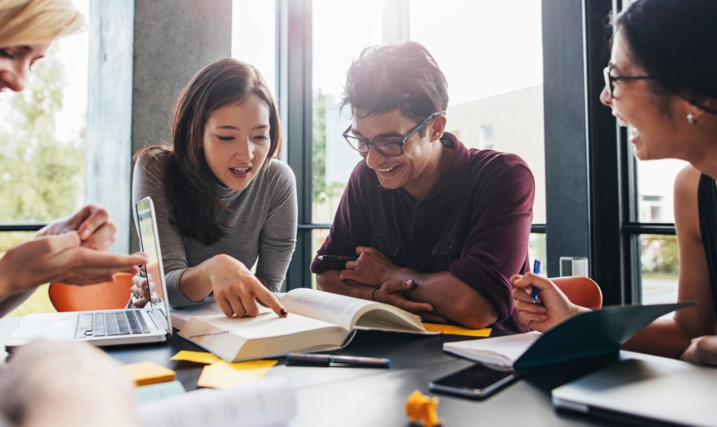 Young students sitting at a table in a library and reading books for their class assignment. University students doing group study in library.