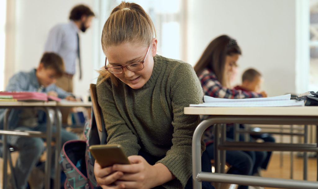 Smiling elementary student using mobile phone while having a class with her friends at elementary school